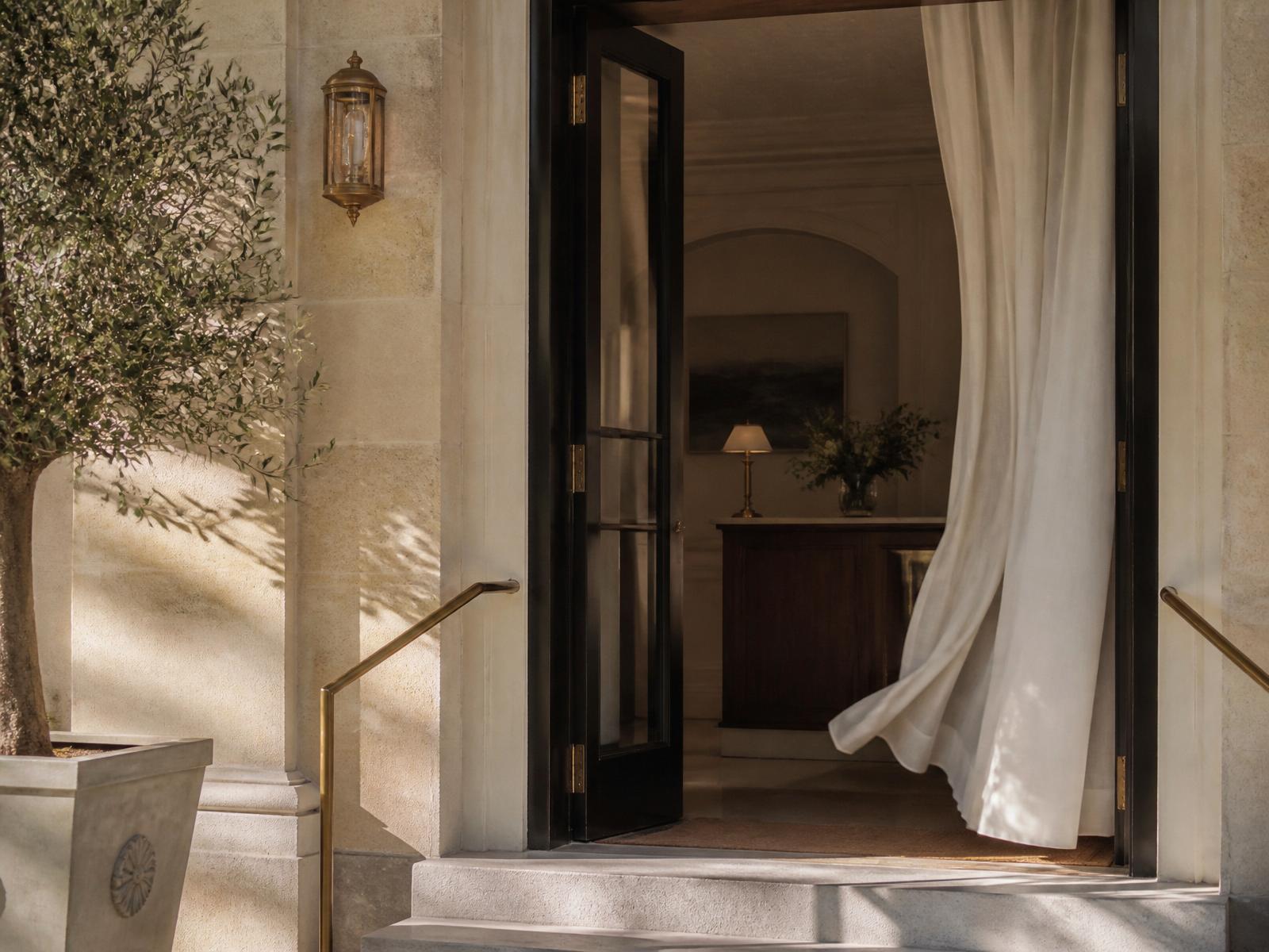 A quiet Mediterranean maison entrance in soft morning light, a linen curtain drifting through an open doorway